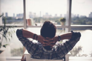 rear-view-of-man-leaning-back-in-chair-with-hands-behind-head-looking-view-through-floor-to-ceiling-windows