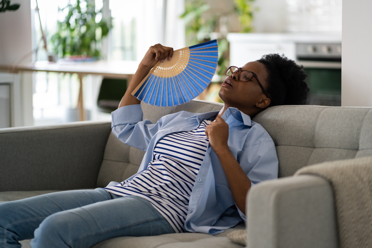 Tired exhausted African American woman sits on sofa at home using paper fans and closing eyes suffering from stuffiness
